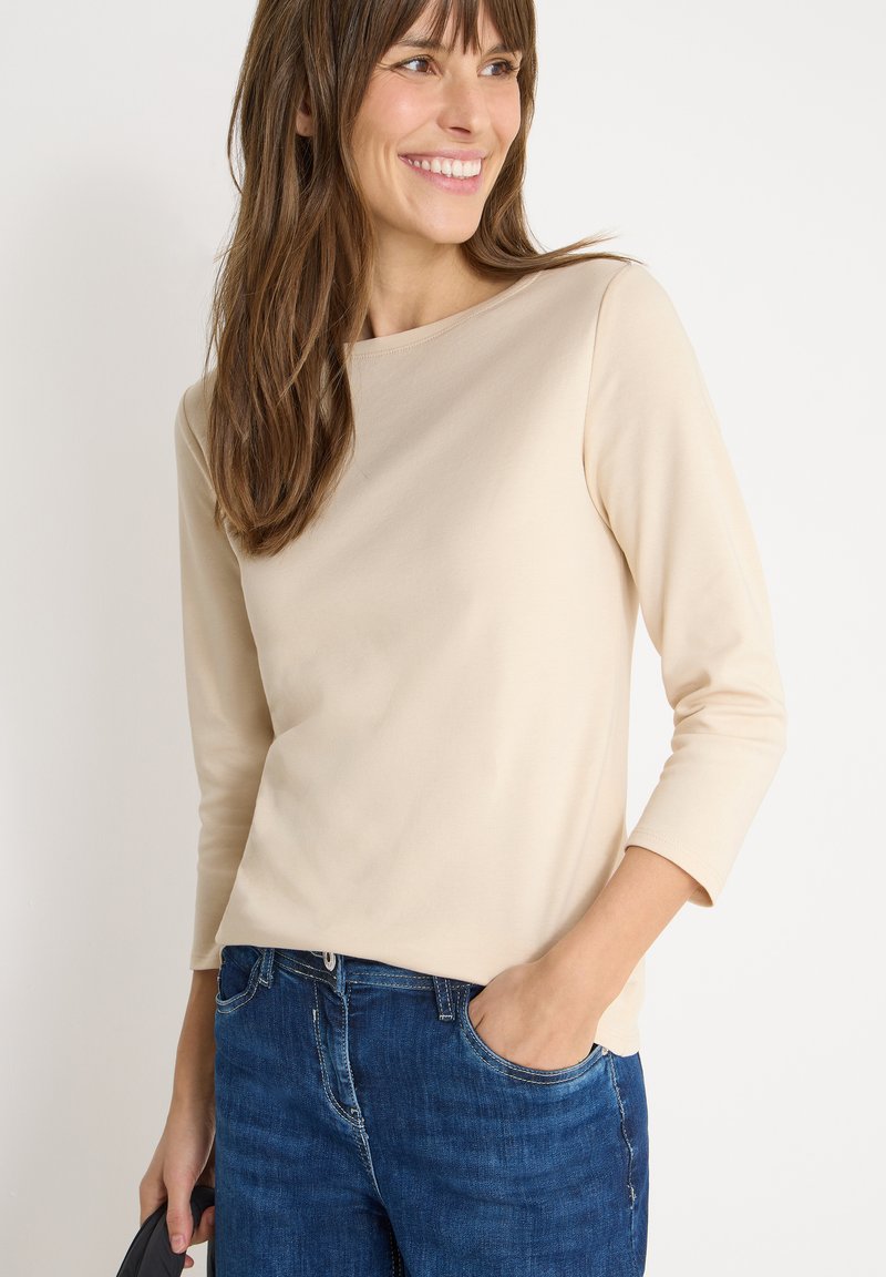 Beige long-sleeve top with a round neckline, made of smooth fabric, paired with blue denim jeans. Model stands with a hand in pocket.