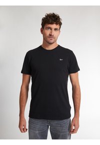 T-shirt nera in cotone con maniche corte e scollatura rotonda, caratterizzata da un piccolo logo bianco sul petto. Indossata con jeans grigi chiari.