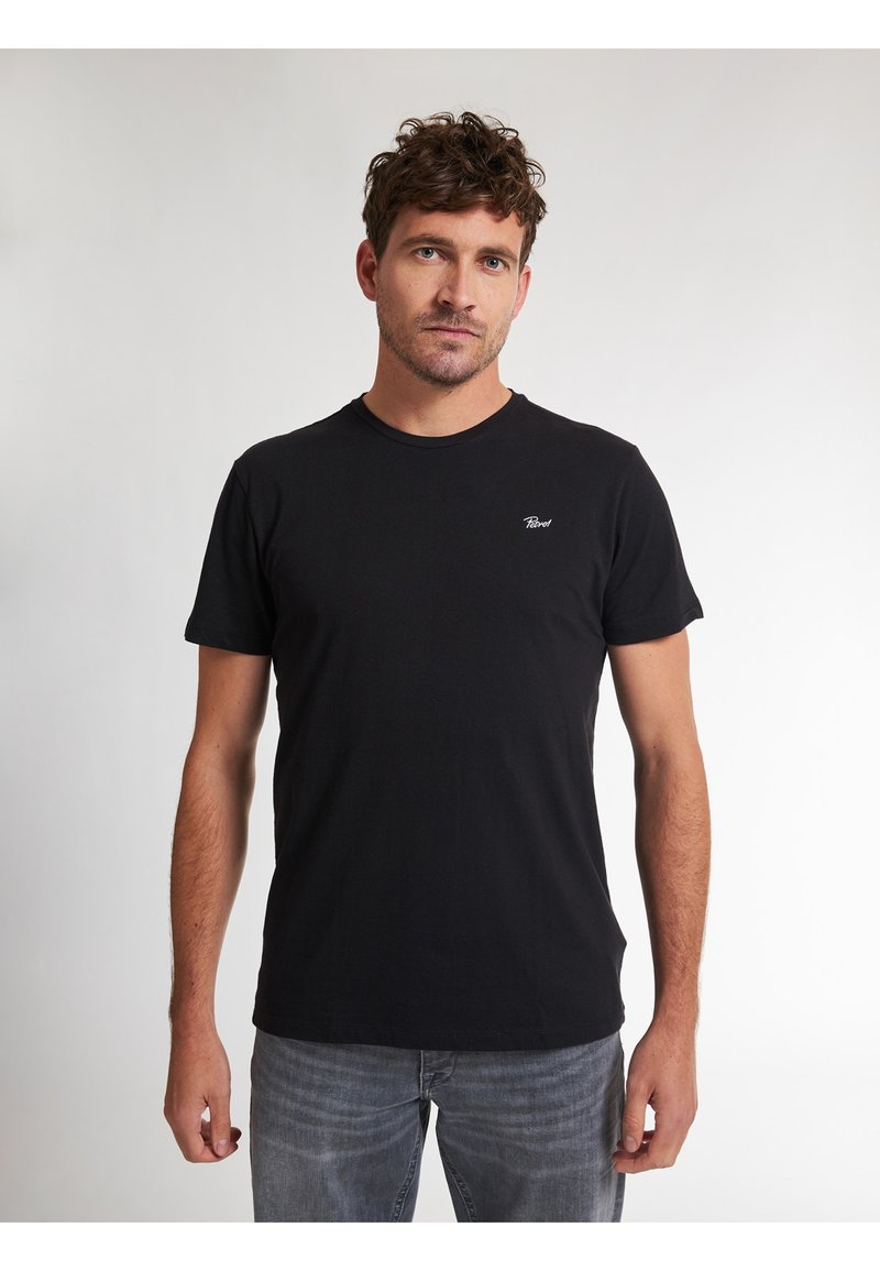 T-shirt nera in cotone con maniche corte e scollatura rotonda, caratterizzata da un piccolo logo bianco sul petto. Indossata con jeans grigi chiari.