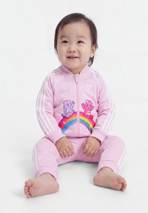 Tout-petit assis pieds nus portant un survêtement rose avec des ours dessinés et un arc-en-ciel sur la veste, souriant et regardant légèrement vers le haut.