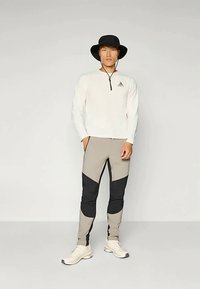 Dugi rukav, bijela majica sa zatvaračem i malim logom; sivo-crne tapered hlače s dizajnom u više boja; osoba nosi crni šešir sa širokim obodom.