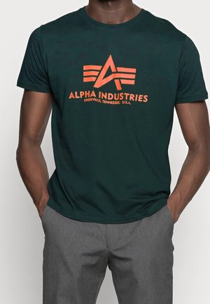 Ciemnozielona bawełniana koszulka z pomarańczowym nadrukiem logo z napisem "ALPHA INDUSTRIES" i trójkątnym wzorem. Krótkie rękawy, okrągły dekolt.