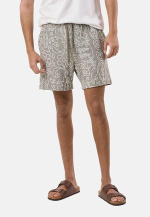 Homme portant un short gris à motifs avec cordon de serrage, une chemise blanche et des sandales en cuir marron à boucle, debout sur un fond neutre.