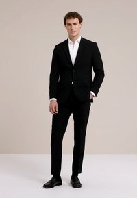 Mladić stoji noseći crno odijelo s bijelom košuljom i crnim elegantnim cipelama na jednostavnoj bež pozadini.