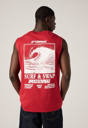 Homme portant un débardeur rouge avec un graphisme vague blanc et le texte « Surf & Swap Invitational » au dos, debout devant un fond uni.