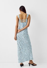 Svetle modré květinové maxi šaty s krajkovým zapínáním na zádech, širokými ramínky a lehkým materiálem, doplněné černými sandály na podpatku.