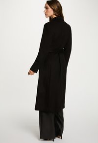Vrouw in een zwarte lang gelaagde jas met riem en zwarte brede broek, staand en kijkend naar de zijkant tegen een effen lichte achtergrond.