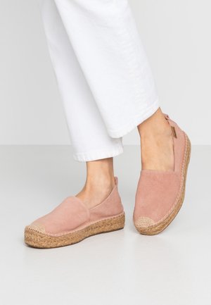 Persoon die witte broek en roze suède espadrilles draagt met geweven jute plateauzolen op een lichte neutrale vloer.