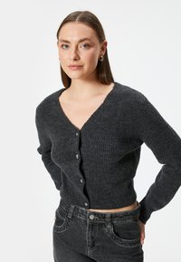 Donkergrijze geribbelde cardigan met een V-hals, voorzien van vier knoopsluitingen, lange mouwen en een getailleerd ontwerp. Draag het over zwarte jeans.