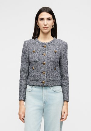 Kvinde med glat mørkt hår iført tekstureret marineblå jakke med guldknapper og lyseblå jeans med høj talje, står mod en ensfarvet baggrund.