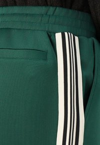 Pantaloni sportivi verzi cu un panou lateral în dungi alb-negru, material texturat și un buzunar discret la spate. Bandă elastică vizibilă la talie.