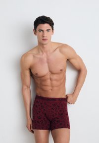 Boxer shorts in burgundy with a black floral pattern. Soft fabric, elastic waistband labeled "BJÖRN BORG," fitted design. Model poses against a plain background.