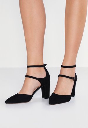 Femme portant des chaussures à talons hauts en daim noir avec des sangles de cheville, debout sur un sol blanc contre un fond blanc uni.
