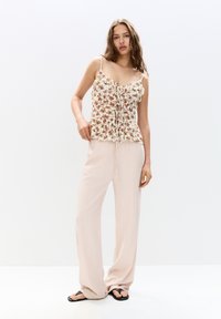 Bluse mit floralem Muster in crème mit Rüschendetails und Spaghettiträgern, kombiniert mit leichten, locker sitzenden beigen Hosen und schwarzen Sandalen.