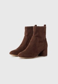 O pereche de botine din suede maro, cu toc pătrat și fermoare pe lateral, prezentate pe un fundal deschis, simplu.