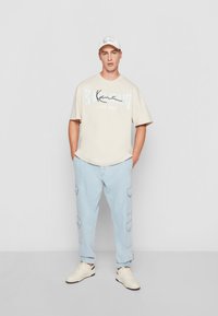T-shirt graphique beige avec le texte "KALANI", pantalon cargo bleu clair et baskets blanches. Comprend également une casquette blanche avec un logo.