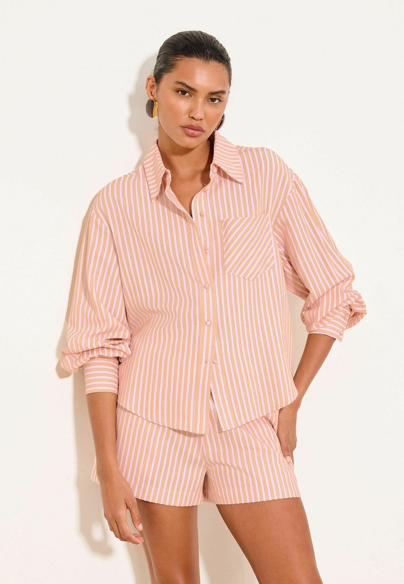 Woman wearing pink and white vertical striped button-up shirt with rolled sleeves and matching shorts, standing against a light background.
