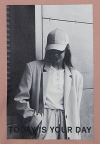 Camiseta rosa claro con un diseño impreso en monocromo de una figura con chaqueta y gorra, con el texto "HOY ES TU DÍA" debajo.