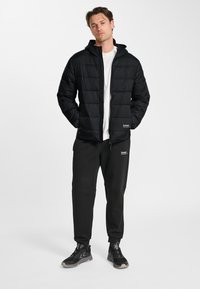 Man staat met handen in de zakken, draagt een zwarte donsjas, een wit T-shirt, zwarte joggingsbroek en zwarte sneakers tegen een effen witte achtergrond.