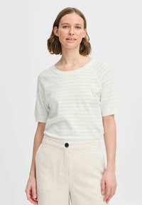 Licht ivoorkleurige gebreide trui met korte mouwen, voorzien van een textuurpatroon van kleine gaatjes. Gepaard met beige linnen broek, knoopdetail zichtbaar.