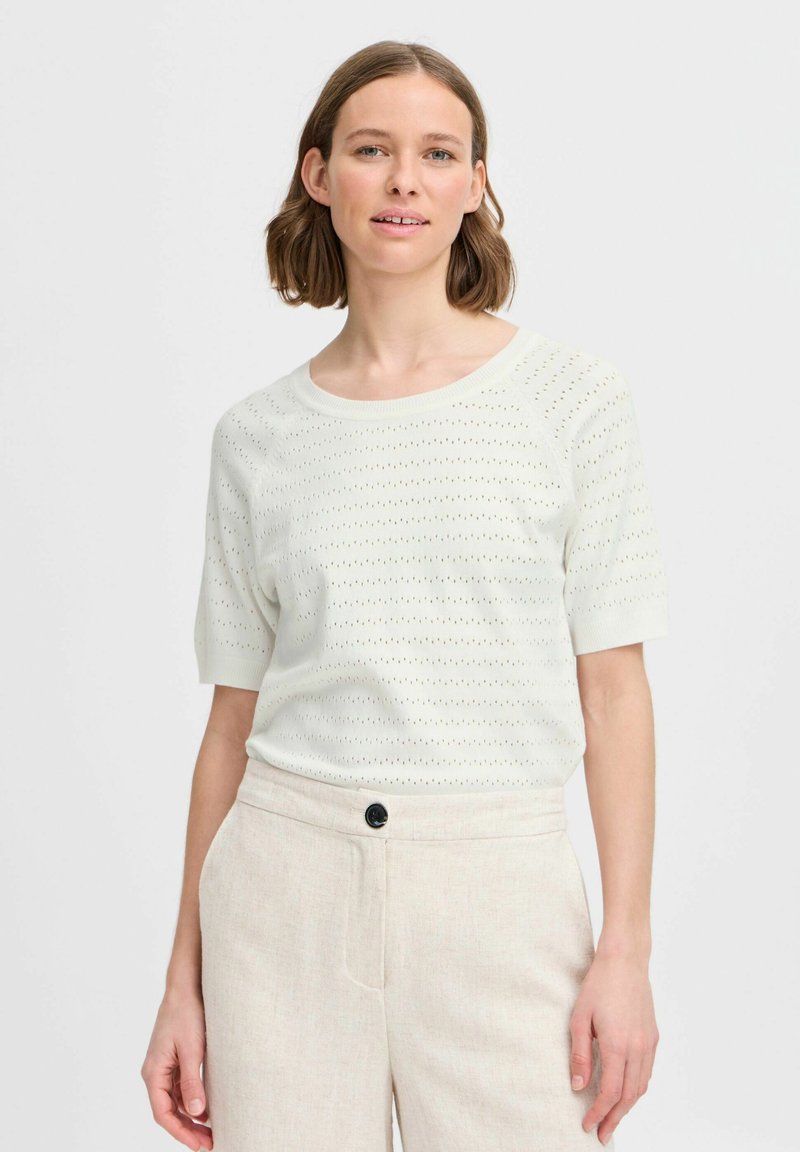 Licht ivoorkleurige gebreide trui met korte mouwen, voorzien van een textuurpatroon van kleine gaatjes. Gepaard met beige linnen broek, knoopdetail zichtbaar.