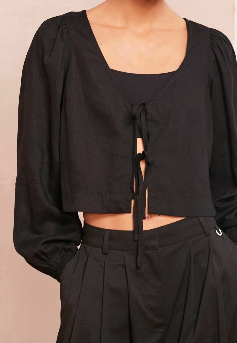 Haut noir court avec des manches longues bouffantes et des liens à l'avant, porté avec un pantalon noir plissé taille haute.