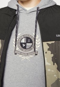 Chaqueta negra y verde oliva con un patrón de camuflaje. Sudadera gris con capucha que presenta un logo circular bordado con texto y detalles de laurel.