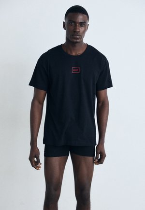 T-shirt in cotone nero con logo quadrato rosso che riporta la scritta "HUGO" sul petto. Il modello indossa slip neri. Design semplice, vestibilità regolare.