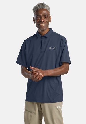 Uomo anziano con capelli grigi sorride, indossa una polo blu navy Jack Wolfskin e pantaloni beige Jack Wolfskin, mani intrecciate.