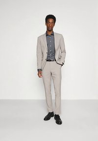 Lichtgrijs pak met een getailleerd ontwerp, gedragen over een donkerblauw overhemd. Soepele textuur, gecombineerd met zwarte schoenen en een riem. Staande houding.