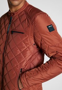 Gesteppte braune Jacke mit Stehkragen, Frontreißverschluss, diagonaler Tasche und schwarzem Logo-Patch am Ärmel. Glatte, glänzende Textur.