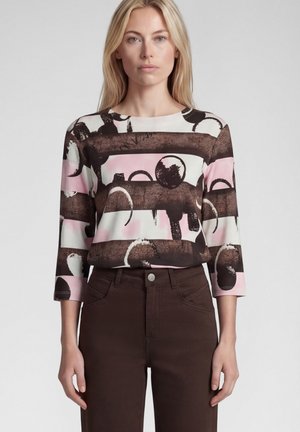 Blonde woman wearing a long-sleeve top with abstract brown, pink, and white patterns, paired with high-waist dark brown pants.