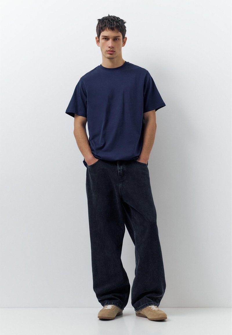 Jeune homme debout les mains dans les poches, portant un t-shirt bleu marine, un jean noir ample et des baskets beiges, devant un mur blanc.