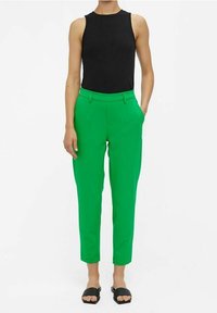 Débardeur noir côtelé associé à un pantalon vert vif fuselé, avec des poches latérales et une texture lisse. Porté avec des sandales noires.