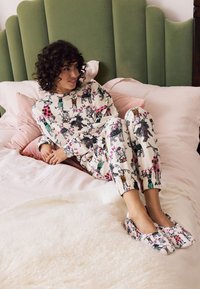 Kreemikas lillemustriga vabaajarõivaste komplekt, millel on elastsed varrukaotsad ja lõdvestunud istuvus. Sobitatud samas stiilis libisemisjalanõudega, asetatud tekstuurse valge tekile.