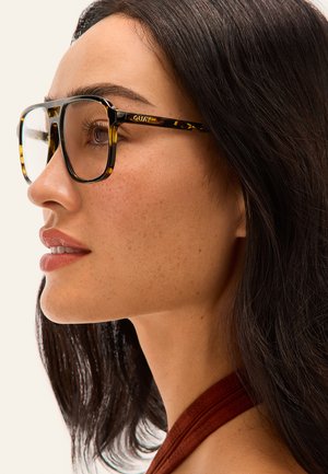 Jeune femme aux cheveux foncés mi-longs portant de grandes lunettes écaille de tortue et un haut de couleur rouille, vue de profil sur un fond clair.