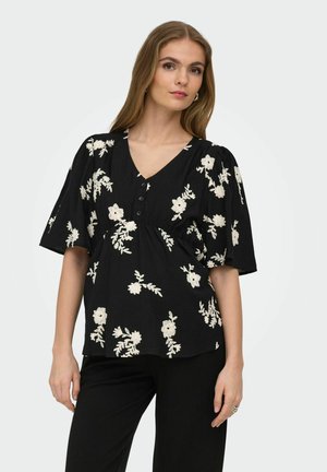 Žena nosiaca čiernu blúzku s bielym kvetinovým vyšívaním, výstrihom do V, gombíkmi a širokými krátkymi rukávmi, v kombinácii s čiernymi nohavicami na jednoduchom pozadí.