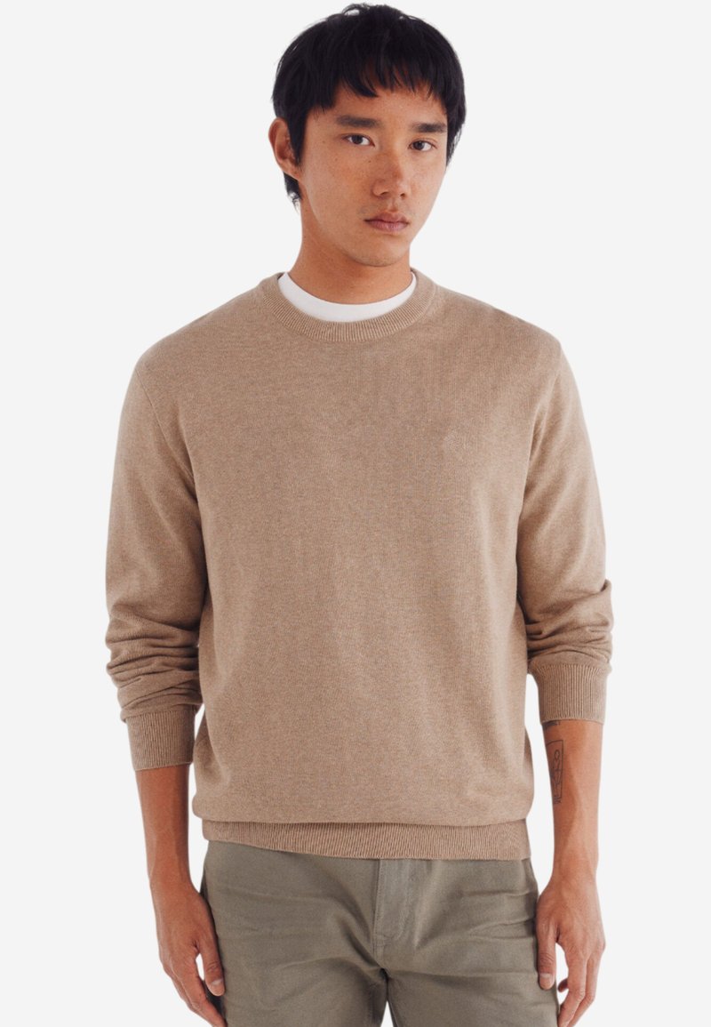 Maglione a maglia beige con scollo rotondo, polsini e orlo a coste. Indossato sopra una T-shirt bianca. Il modello ha i capelli corti e scuri e indossa pantaloni color verde oliva.