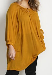 Mosterdgele long-sleeve tuniek met een keyhole-detail bij de nek en elastische mouwen. Gemaakt van een lichte, gestructureerde stof.