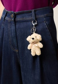 Pequeño llavero de oso de peluche beige con pelo texturizado y un lazo, sujeto a un bolsillo de mezclilla azul oscuro.