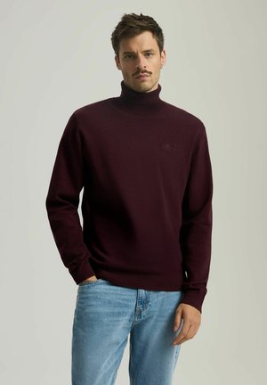 Bordowy sweter z golfem wykonany z dzianiny, z długimi rękawami i subtelnym logo na piersi, zestawiony z jasnoniebieskimi dżinsami.