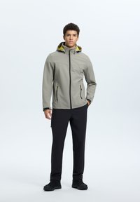 Veste d'extérieur gris clair au design ajusté, avec des zippers noirs et une capuche intérieure jaune, associée à un pantalon noir et des chaussures noires.