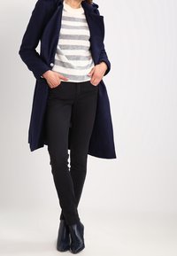 Marinblå trenchcoat över en grå och vitrandig tröja, svarta figursydda byxor och marinblå ankelskor, vilket framhäver en skräddarsydd silhuett och texturer.