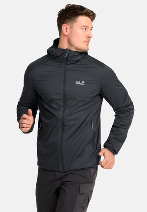 Man draagt een zwarte Jack Wolfskin capuchonjas en donkere broek, gepositioneerd alsof hij aan het joggen of rennen is, kijkt naar zijn rechterkant.