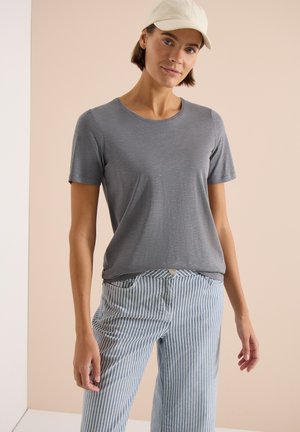 Frau trägt ein graues kurzärmeliges Shirt, blau-weiß gestreifte Hose und eine weiße Kappe, steht vor einem neutralen beigen Hintergrund.