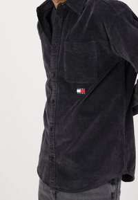 Camicia in velluto a coste grigia scura con bottoni al davanti, una tasca sul petto e un piccolo logo "TOMMY JEANS". La trama presenta una costina verticale.