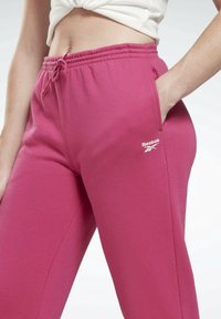 Różowe dresy wykonane z miękkiego materiału, z elastycznym paskiem w talii, sznurkiem do regulacji, bocznymi kieszeniami oraz białym logo Reebok na udzie.