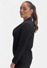 Blazer nero su misura con design aderente, revers a lancia e tessuto liscio. Vista laterale che mostra spalle strutturate e tasche discrete.