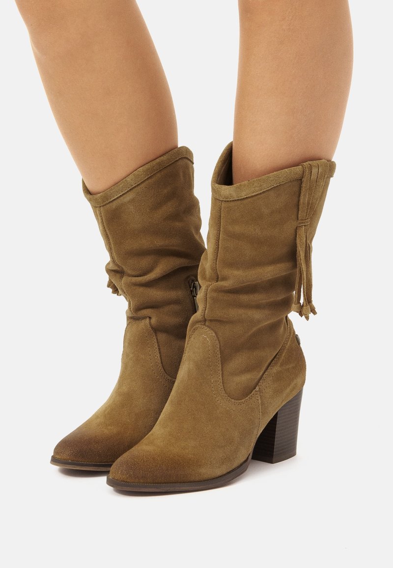 Botas de ante marrón, con un diseño holgado, punta afilada, tacón bloque de altura media y borlas decorativas en el asta. Detalle de cremallera en un lado.