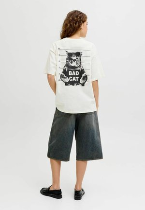 Kvinde iført hvid T-shirt med katte-fængselsfoto, der holder et skilt med "BAD CAT" på ryggen, kombineret med mørke, vidde ben-shorts og sorte sko.